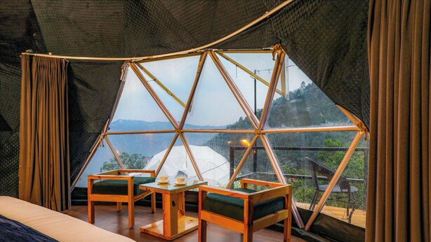 A tented accommodation with a wooden interior, a bed, two chairs, a table, and a view of the outdoors.