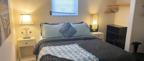 A bedroom with a bed, bedside lamps, a window with blinds, a hanging coat rack, and a small nightstand.