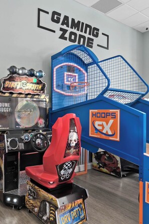 Espaço de jogos com máquinas de fliperama, uma cesta de basquete e uma cadeira gamer vermelha.