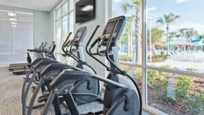 A row of treadmills with a TV mounted on the wall, large windows, and a view of a palm tree-lined outdoor area.