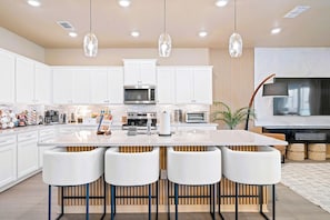 A modern kitchen with white cabinets, a central island with bar stools, a microwave, and a large flat-screen TV. - New Resort, Private Pool & Spa, Outdoor Theater, Game Room, Minutes to Disney World (Clermont)