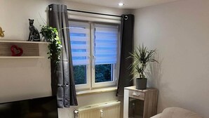 Una sala de estar con una televisión sobre un mueble de madera, un sofá color beige, una mesa de centro, un radiador y una ventana con cortinas.