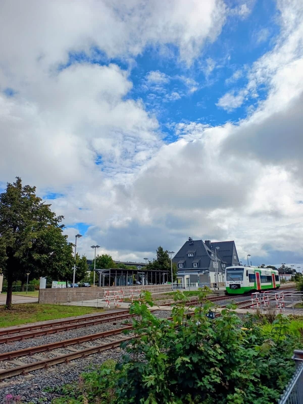 A train station with tracks, a green and white train, and a building with a sloped roof.