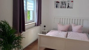 Un dormitorio con cama, ventana con cortinas, radiador, mesita de noche, lámpara y una pared decorada con obras de arte.