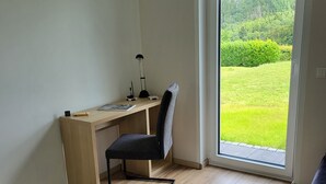 A modern room with a wooden desk, a black office chair, a round coffee table, and a view of a green landscape through a large window. - Vacation home Tannenblick in the heart of the Eifel (Hellenthal)