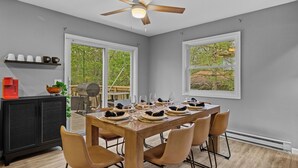 A dining area with a wooden table set for six, surrounded by six chairs. There is a ceiling fan above the table, and a black cabinet with shelves on the left.