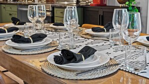 A dining table set with plates, glasses, and cutlery, with a kitchen in the background.