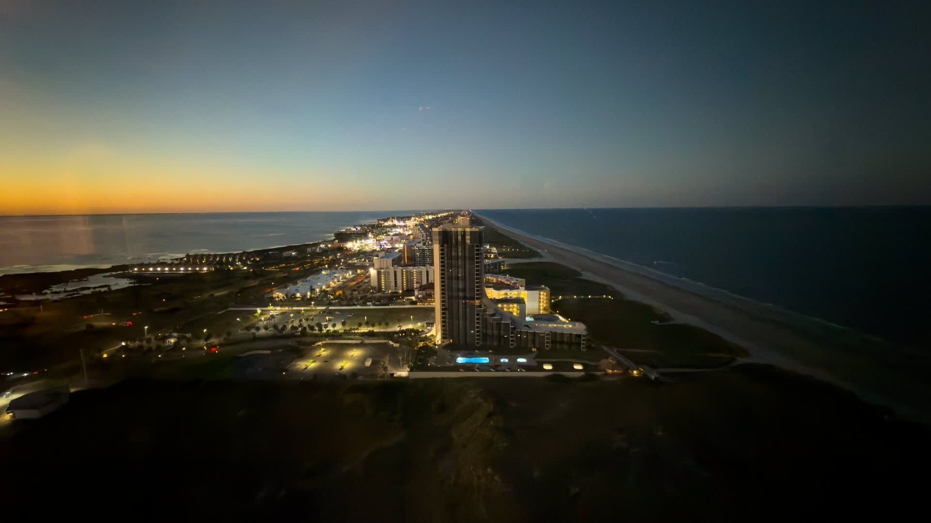 An aerial view of a coastal hotel with multiple buildings, a beachfront, and a well-lit promenade.