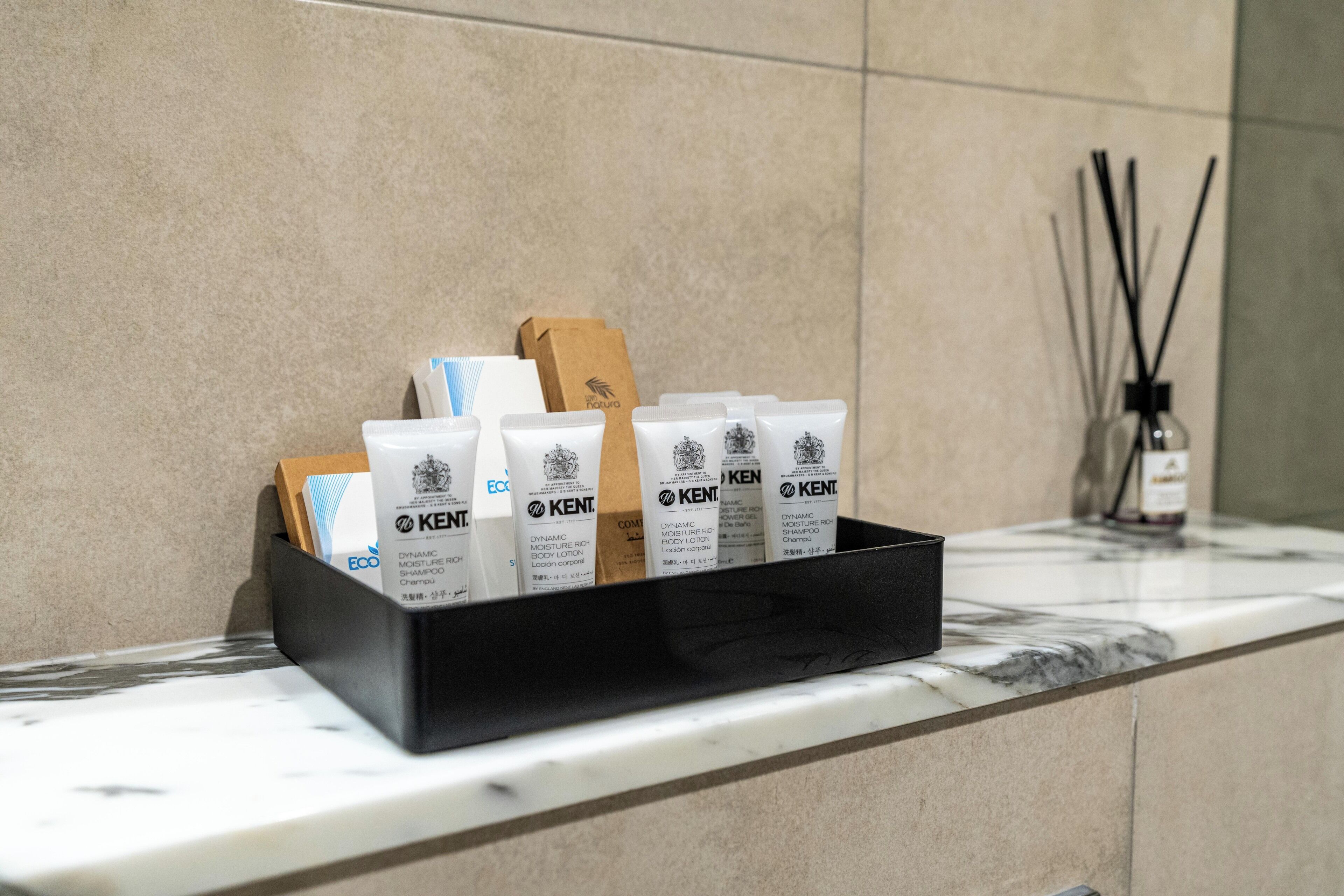 Ein schwarzes Tablett auf einer Marmorarbeitsplatte mit mehreren weißen Röhren der Marke 'KENT', einer braunen Papiertüte und einer kleinen Flasche für einen Reed-Diffuser.