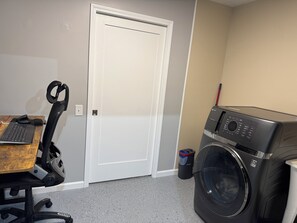 A laundry room with a washing machine, a door, and a desk with a keyboard and mouse.