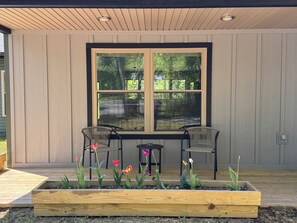 A covered patio with two chairs and a table, surrounded by raised garden beds with flowers.