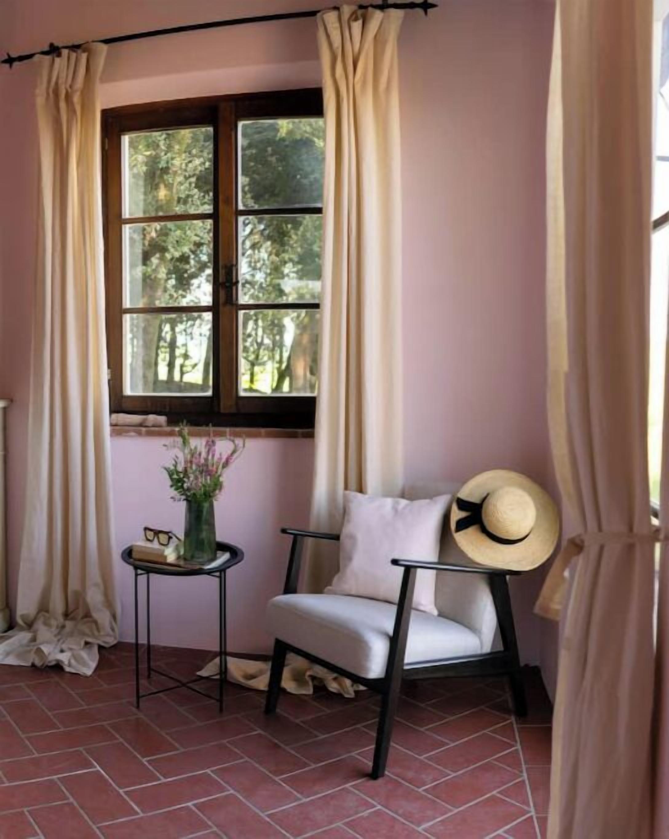 Una habitación de esquina con ventana, una silla, una mesita con un florero y vasos, y cortinas.
