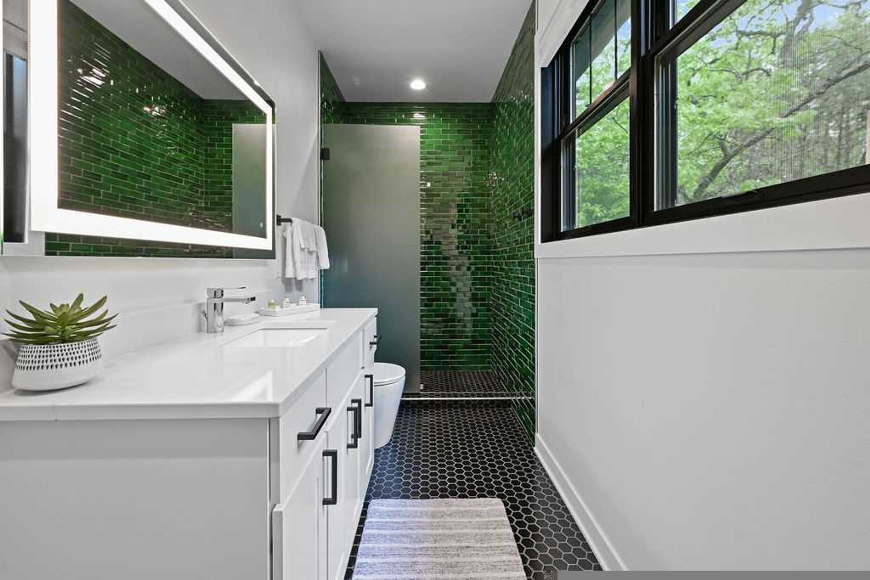 Un baño moderno con un tocador blanco, una zona de ducha con azulejos verdes y una ventana que proporciona luz natural.