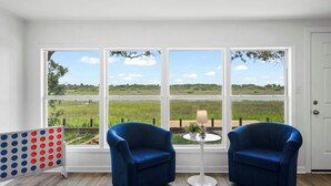 Een kamer met twee blauwe fauteuils, een kleine ronde tafel met een lamp, en uitzicht op een grasveld door grote ramen.