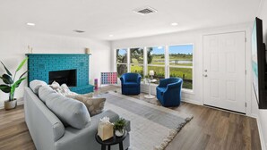 Een woonkamer met een blauwe bakstenen open haard, een grijze bank, twee blauwe fauteuils en een groot raam met uitzicht op een groen landschap.