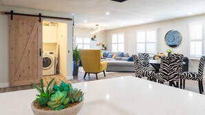 Een moderne woonkamer met een grote witte tafel, een gele fauteuil en stoelen met een zebrapatroon. Er is een houten deur die leidt naar een wasruimte, en een klok aan de muur.