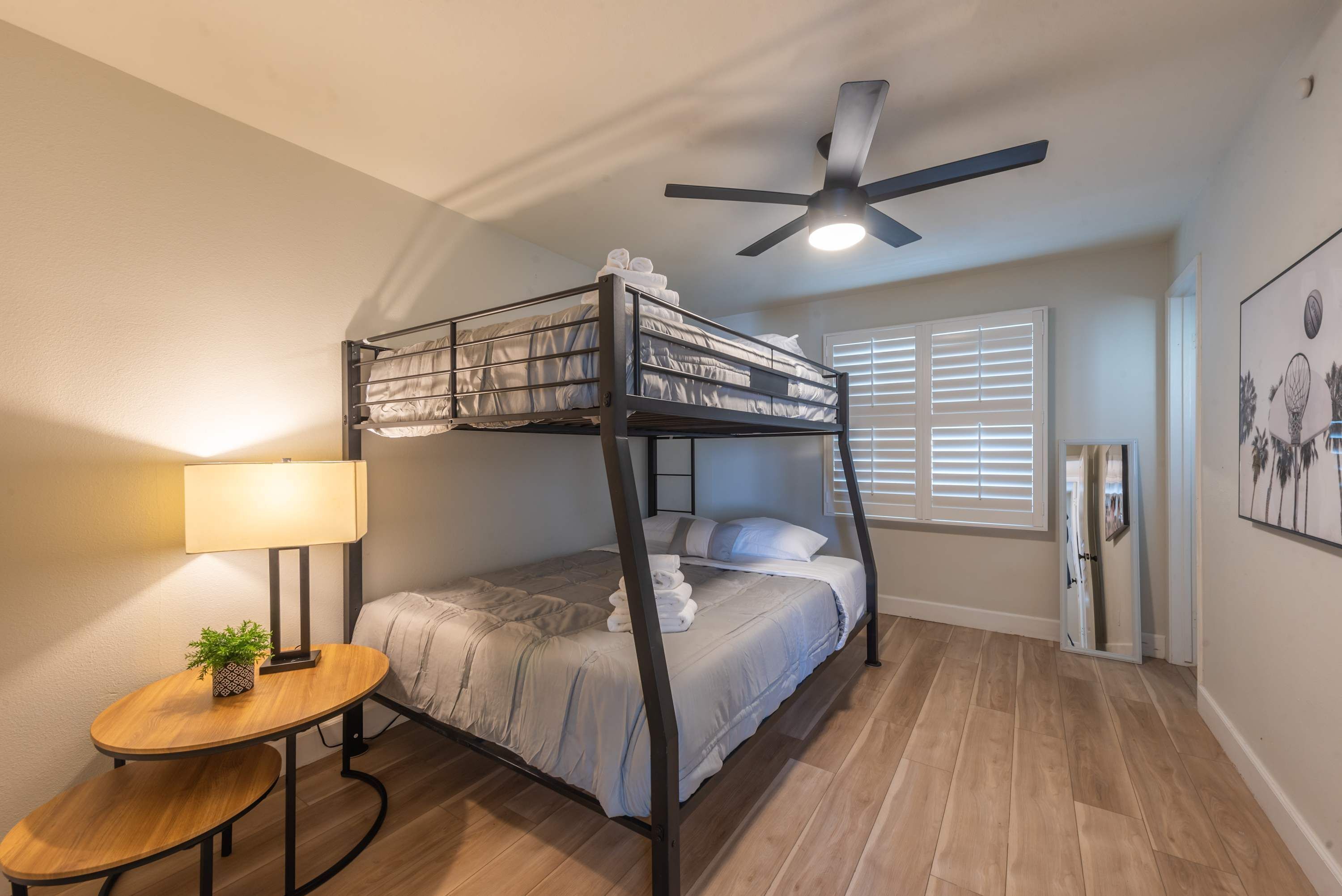 A bunk bed room with a ceiling fan, a bedside table with a lamp, and a window with blinds.