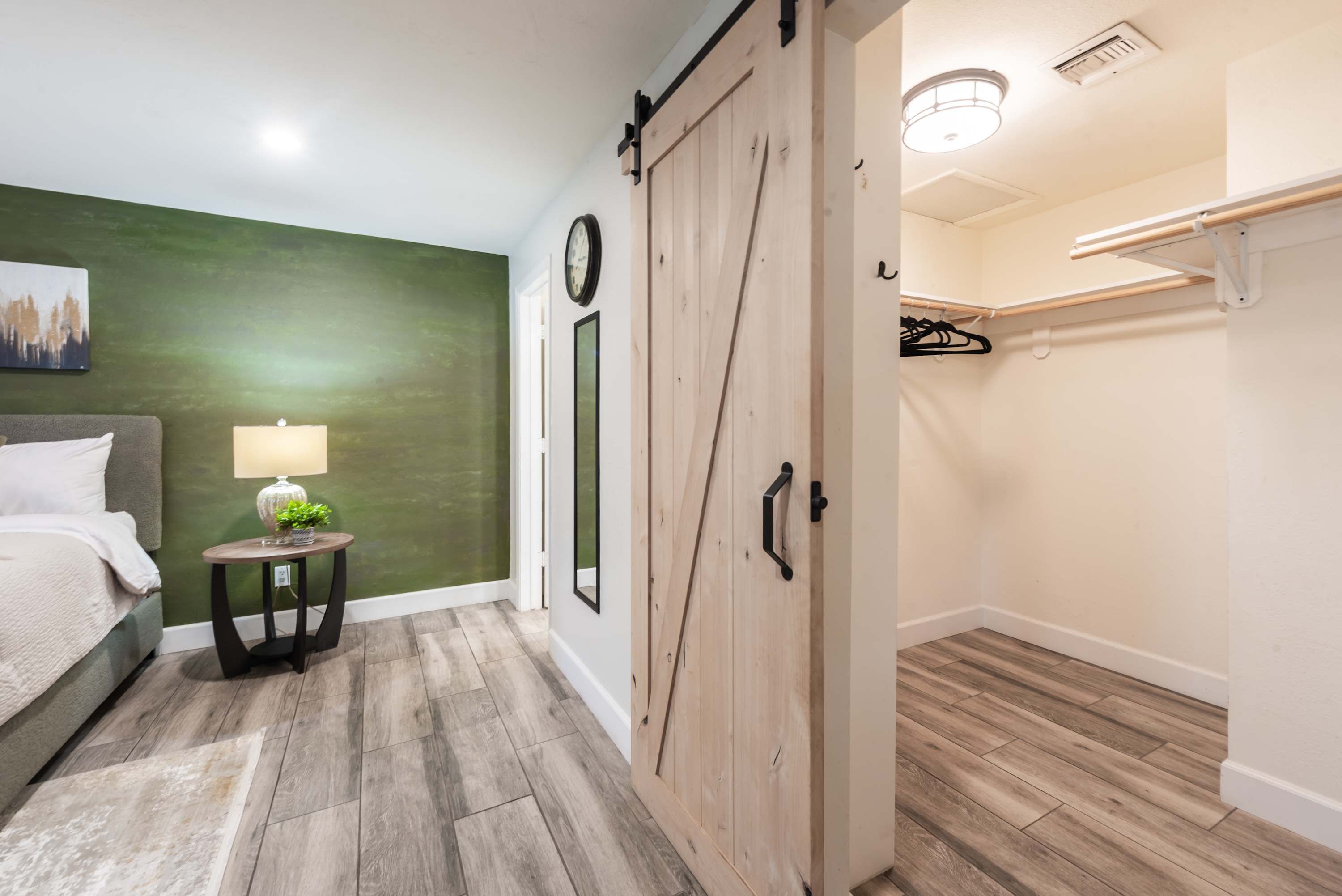 Un dormitorio con cama, mesita de noche con lámpara, pared de acento verde, armario de madera y un pasillo con perchero.