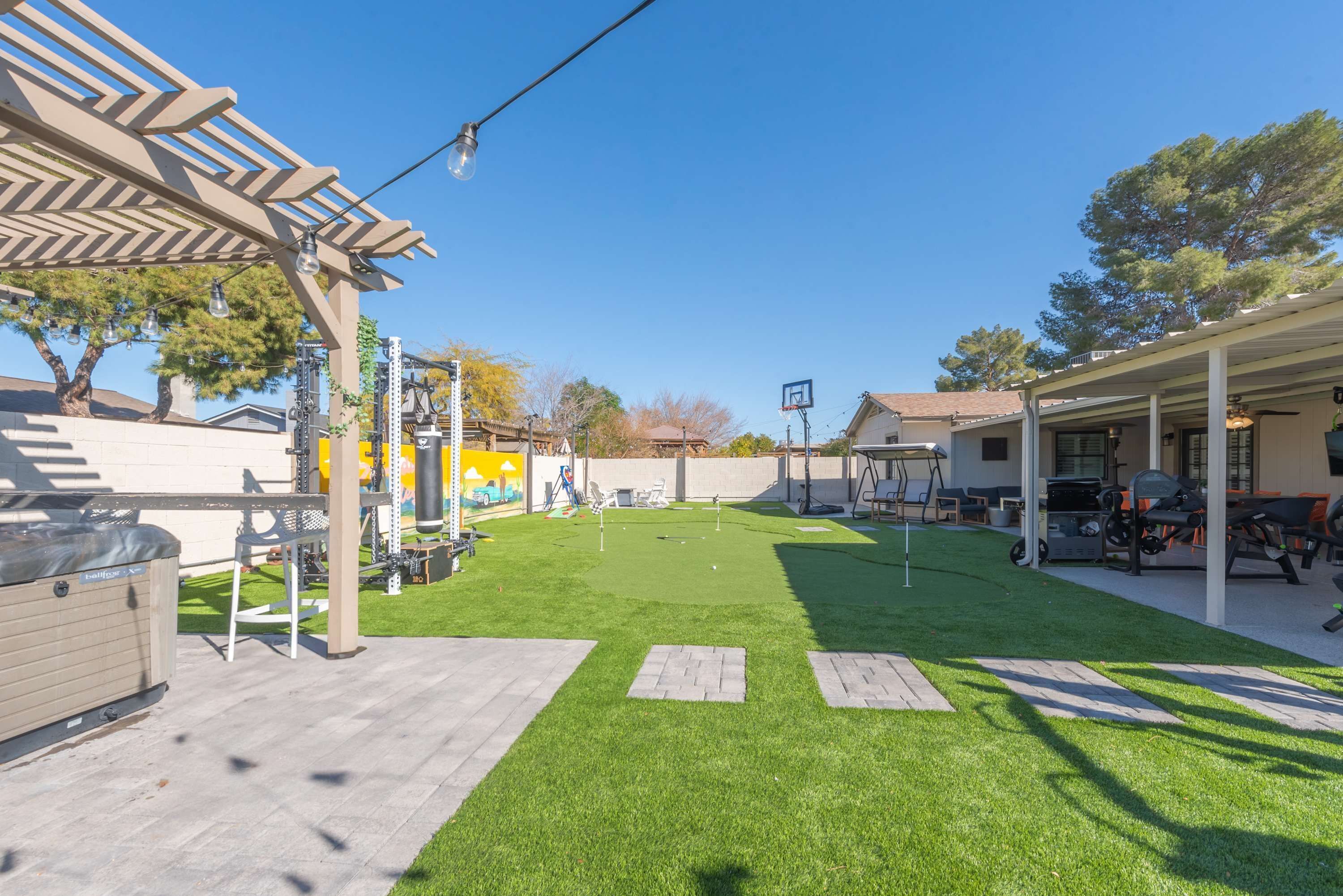 Un patio trasero con césped artificial, una pérgola, mobiliario exterior y un aro de baloncesto.