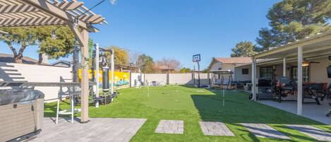 Un patio trasero con césped artificial, una pérgola, mobiliario exterior y un aro de baloncesto.