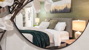 Een slaapkamer met een bed, kussens, een nachtkastje, een lamp en een schilderij aan de muur.