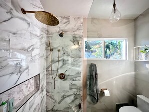 A modern bathroom with a glass shower enclosure, marble walls, a window with a view of greenery, and a towel rack.