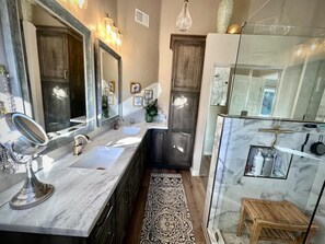 A modern bathroom with a large mirror, two sinks, a shower area, and a wooden stool.