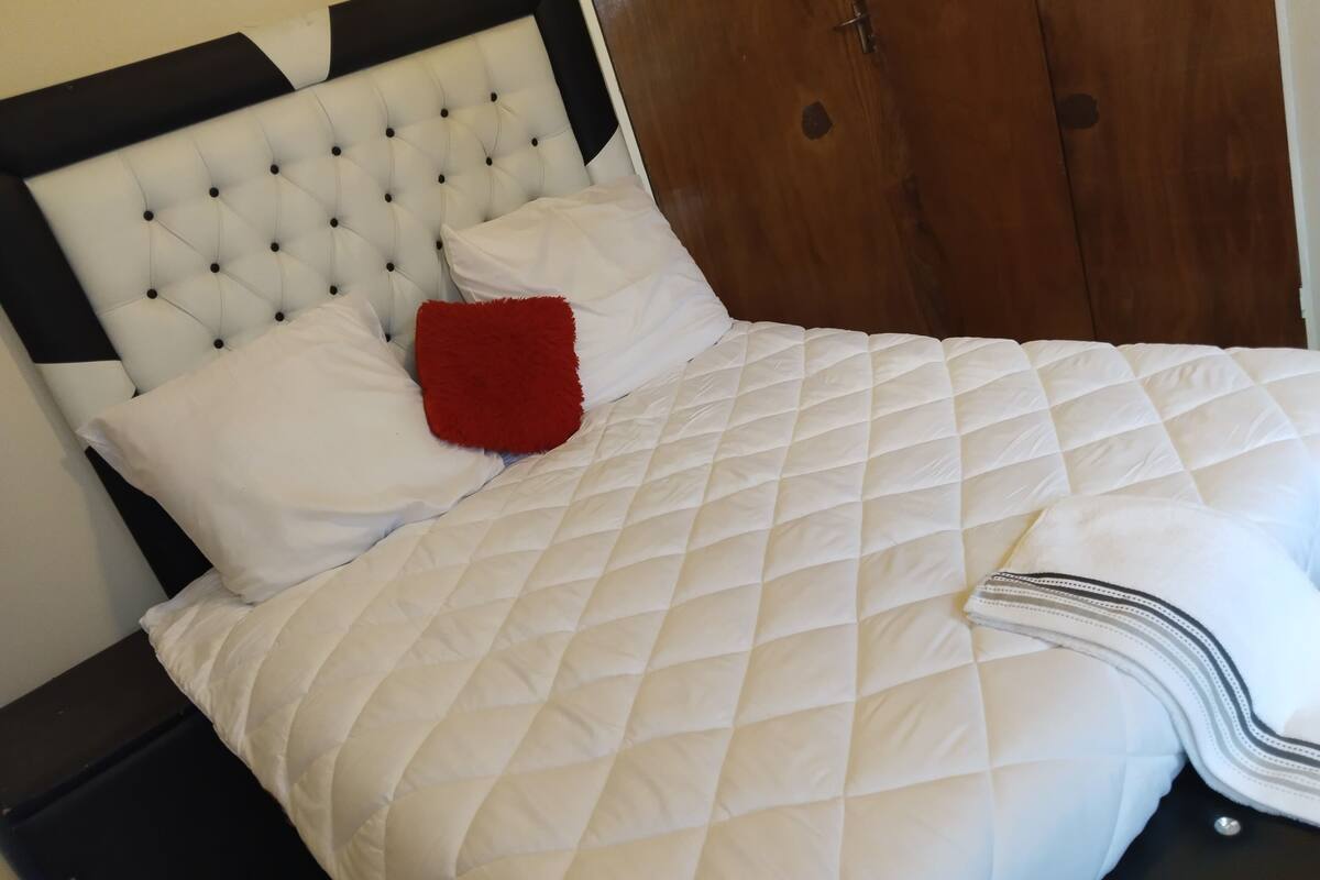 Una cama con edredón blanco acolchado, una almohada roja y un cabecero negro.