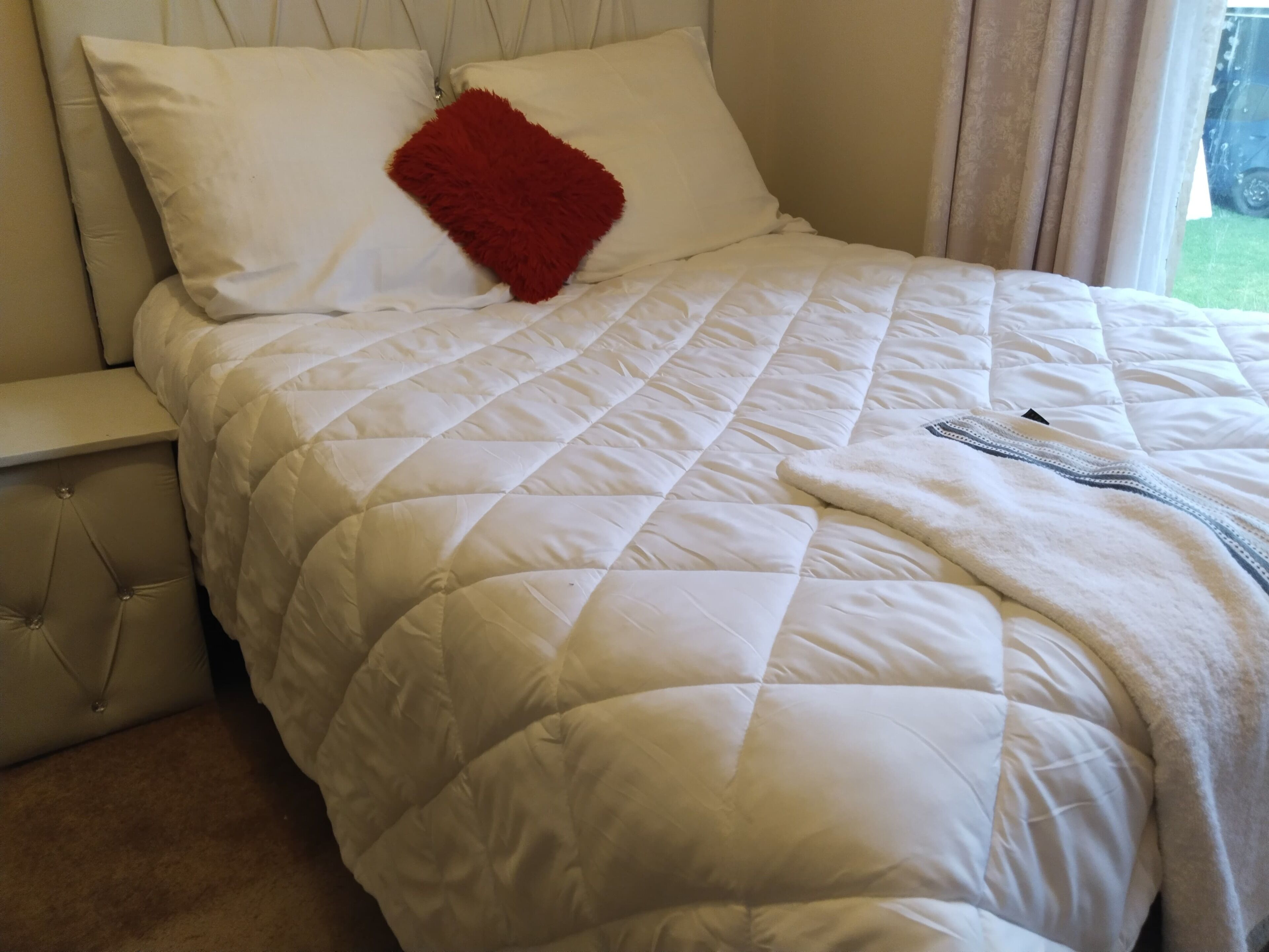 A neatly made bed with a white quilt and a red accent pillow, a tufted headboard, and a window with curtains.
