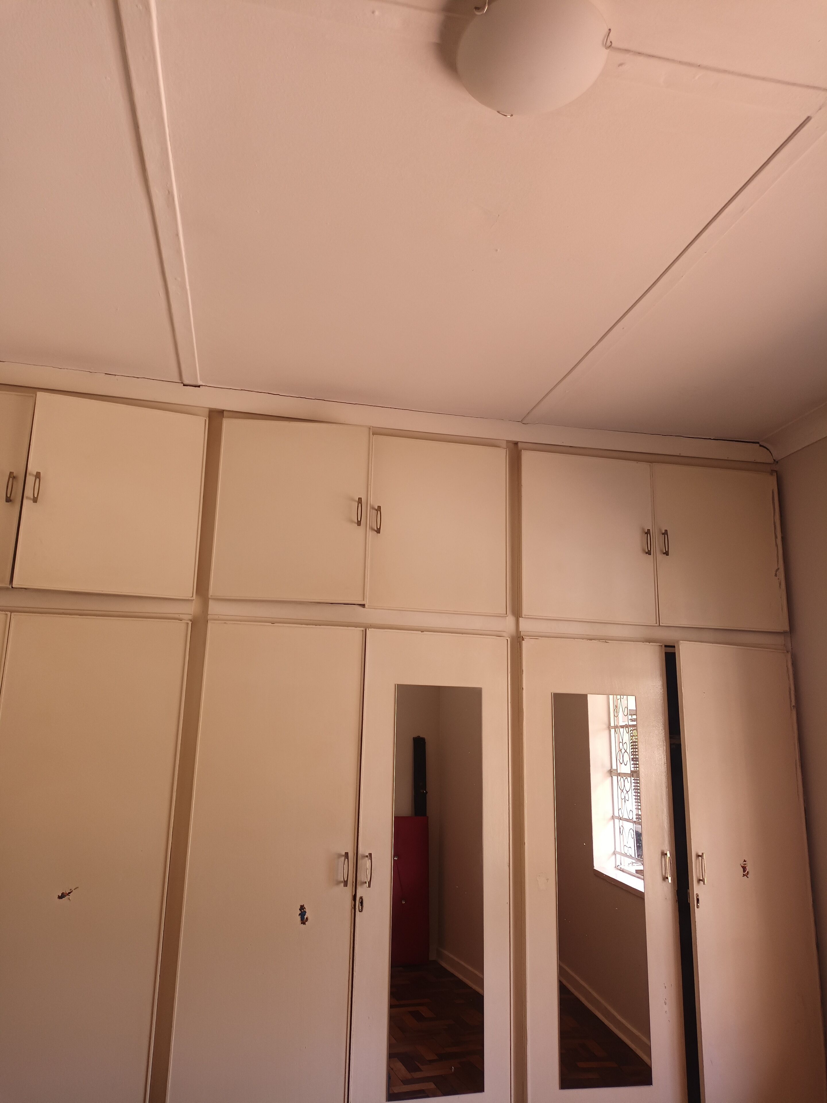 A room with a white ceiling, white walls, and a wooden floor. There are multiple white doors with handles, and a red door is visible in the background.