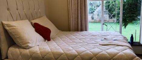 A bed with a quilted headboard, a red pillow, and a window with a view of greenery outside.