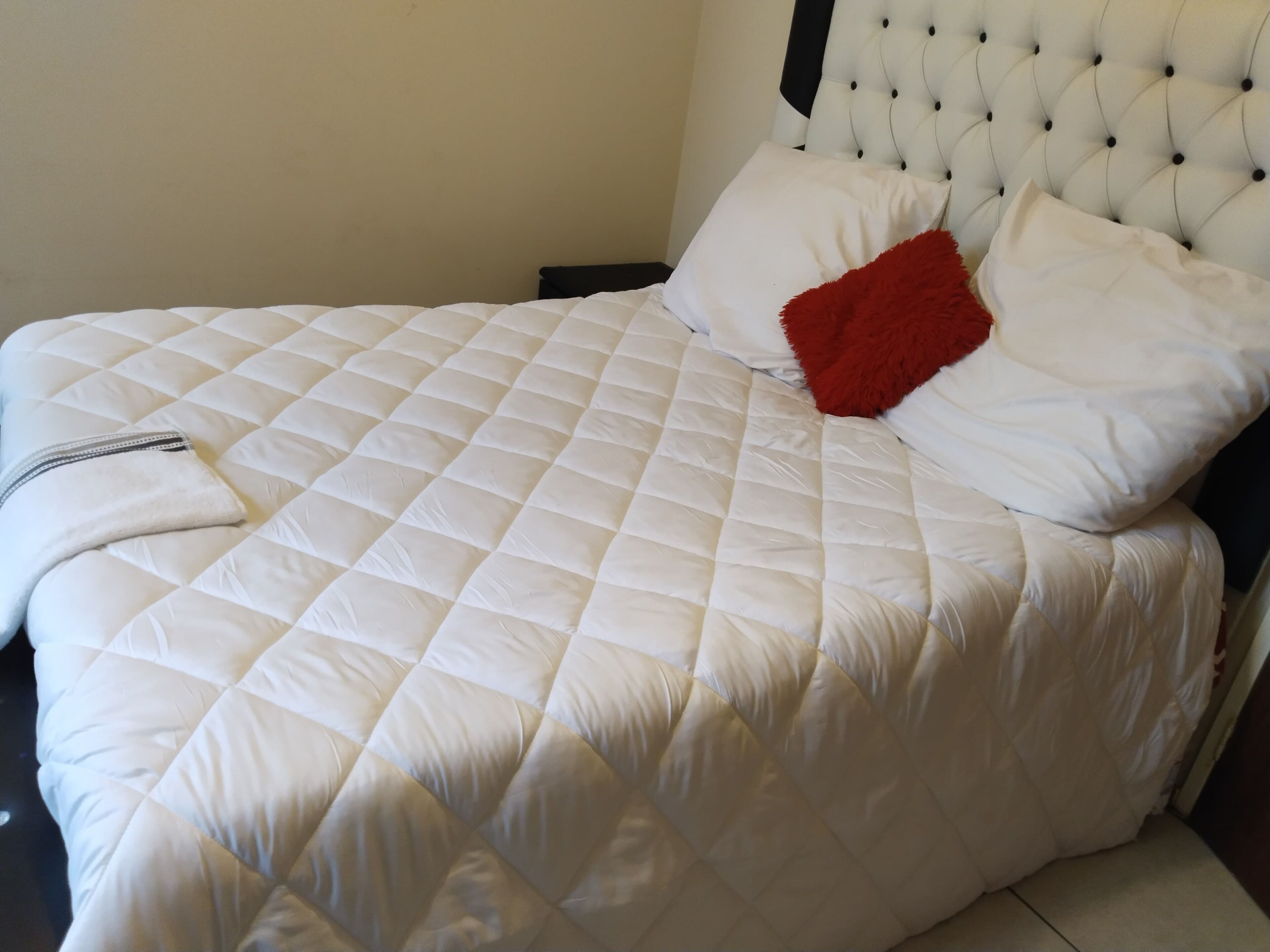 A bed with a quilted white duvet, a red pillow, and a white blanket.