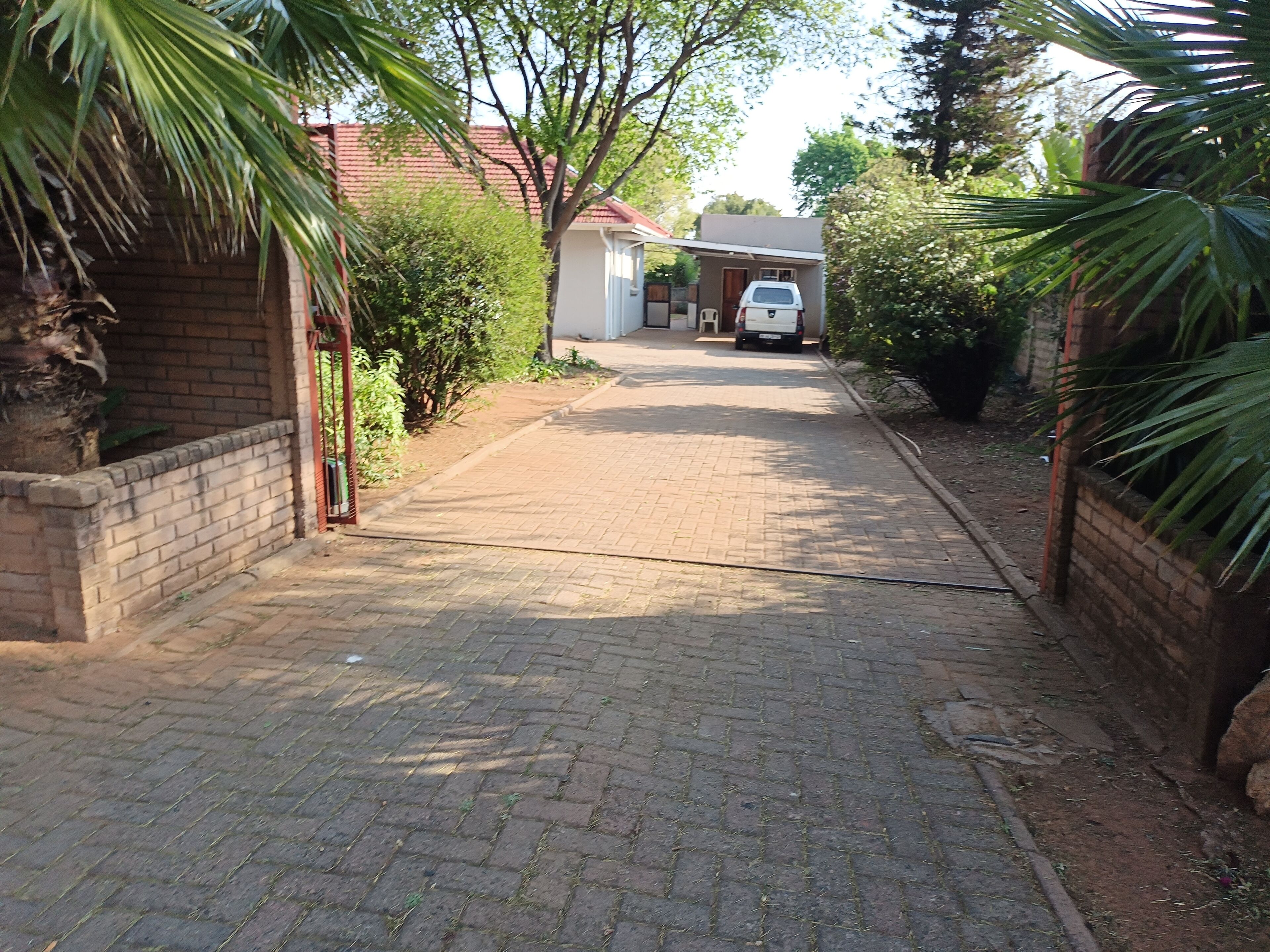 A paved driveway leading to a house with a red roof, surrounded by greenery and a brick wall.