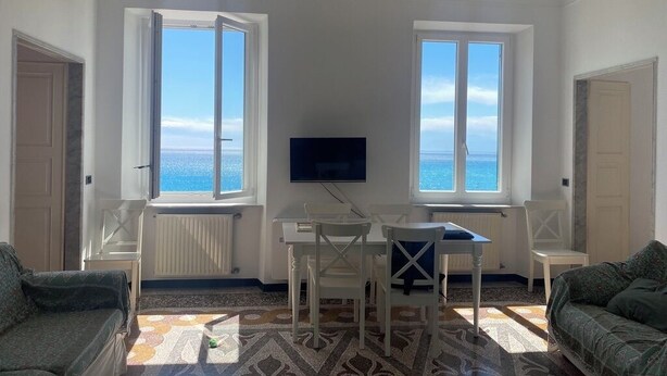 A room with a dining table, two chairs, a TV, and a sofa. There are two windows with a view of the sea. - Green Wave apartment (Camogli)