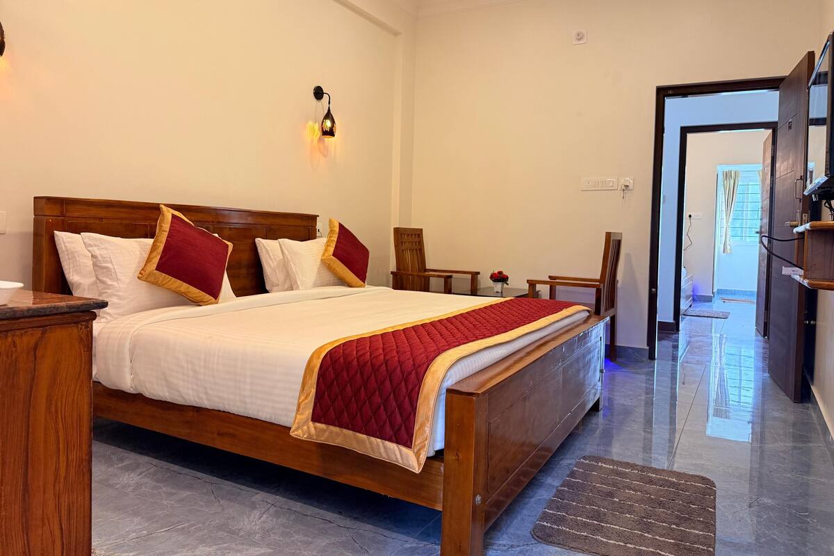 Habitación de hotel con cama de madera, ropa de cama blanca, almohadones rojos, una mesita de noche, una silla, una pequeña mesa con un florero y vista al exterior por una puerta abierta.