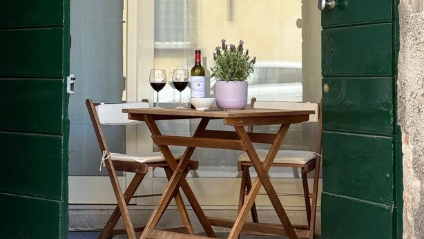A wooden table with two chairs, a potted plant, and two wine glasses with a bottle of wine on a balcony. - Romantic Loft in the historic center of Lucca, ideal for visiting the city on foot (Lucca)