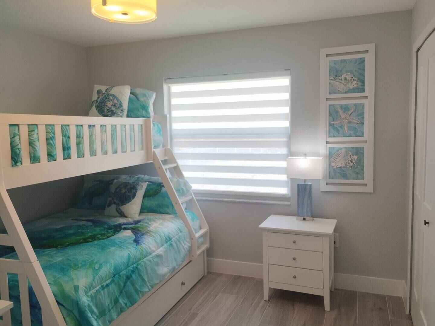 A child's bunk bed with a blue and green ocean-themed bedspread, a small bedside table with a lamp, and framed ocean-themed artwork on the wall.