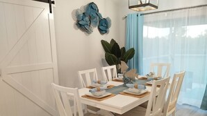 Comedor con una mesa blanca dispuesta para cuatro personas, sillas blancas, un elemento decorativo azul en la pared y una planta grande.