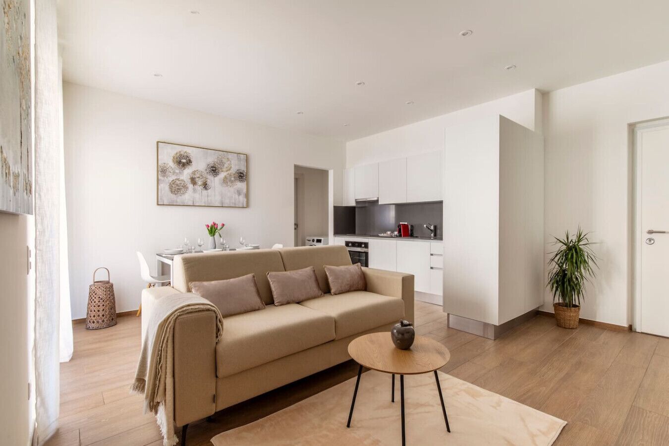 Una sala moderna con un sofá beige, una mesita redonda y una cocina al fondo.