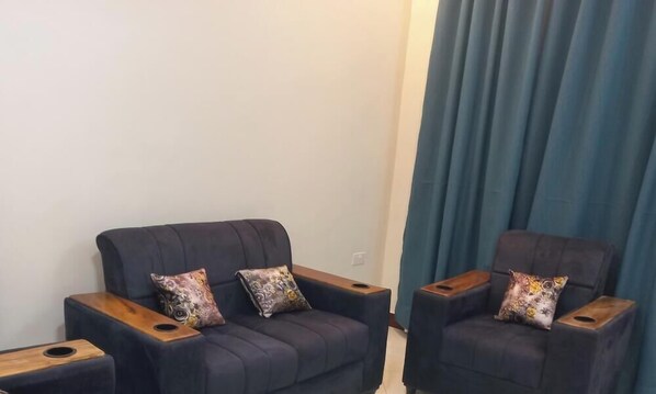 Dos sillones oscuros con mesitas de madera, una pared lisa y una cortina azul.