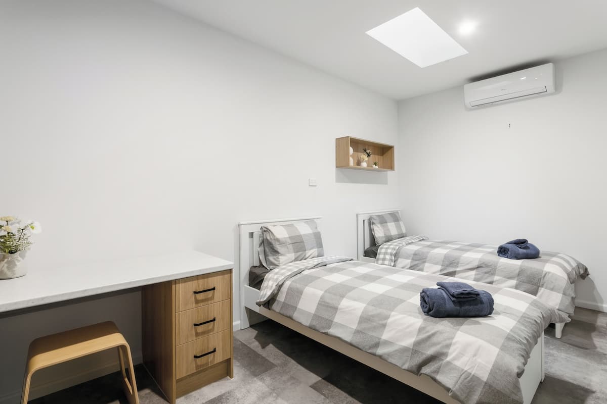 A bedroom with two beds, a desk, a chair, a small shelf, and an air conditioning unit.