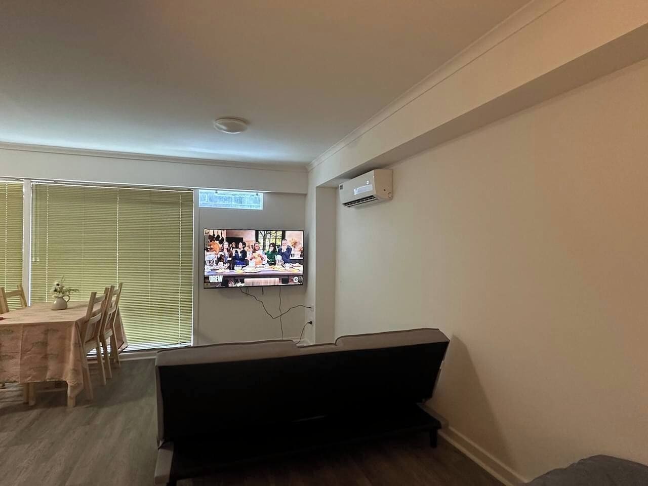 A living room with a TV mounted on the wall, a sofa, a dining table with chairs, and a window with blinds.