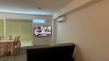 A living room with a TV mounted on the wall, a sofa, a dining table with chairs, and a window with blinds.