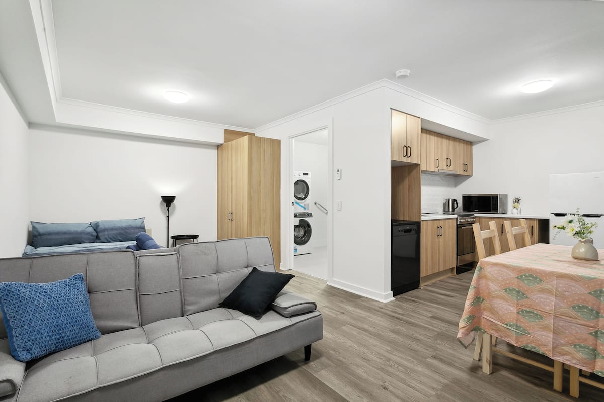 A modern living room with a grey sofa, a dining table with a tablecloth, and a kitchen area with wooden cabinets and appliances.