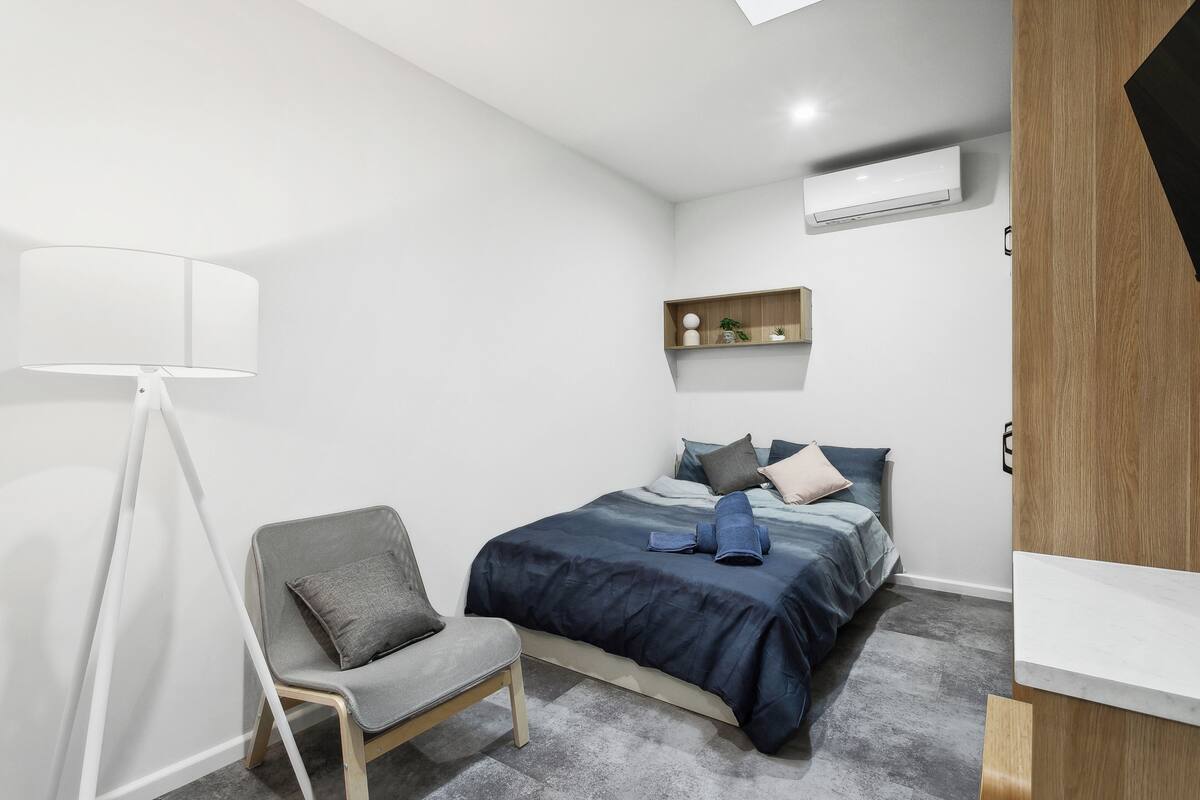 A modern bedroom with a bed, a chair, a shelf with decorative items, and an air conditioning unit.