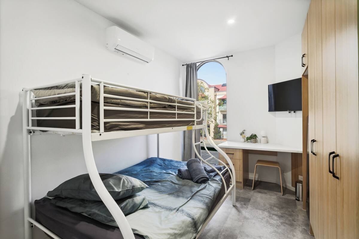 A bunk bed with a desk and chair, a TV mounted on the wall, and a window with a view of buildings outside.