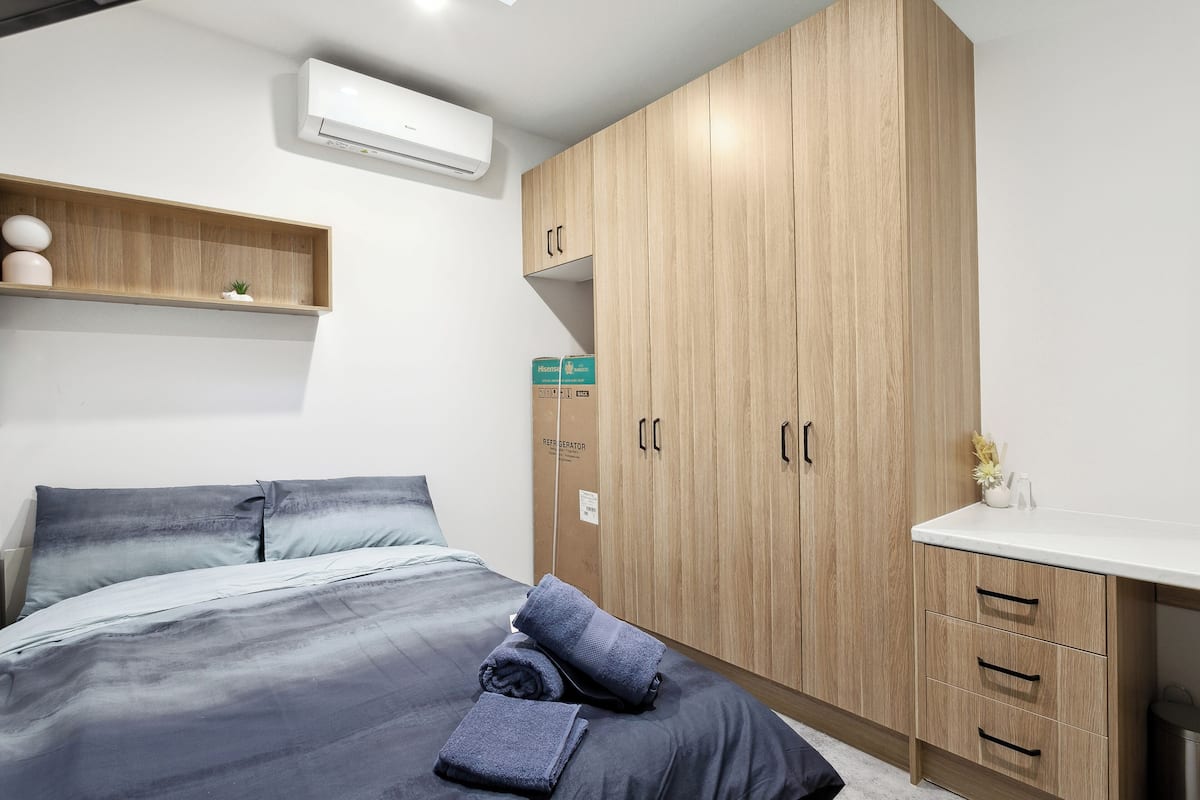 A bedroom with a bed, a wooden wardrobe, a small shelf with decorative items, and a white air conditioning unit.