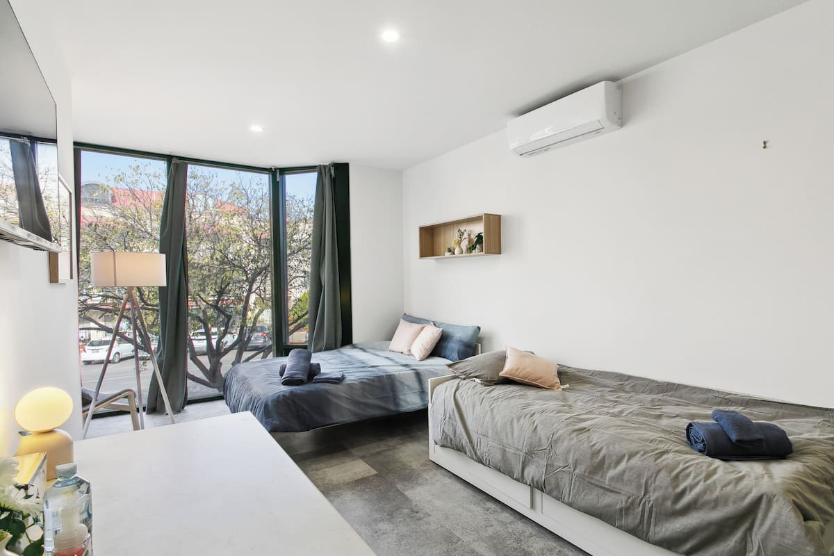 A modern hotel room with two beds, a desk, a chair, and a large window offering a view of trees and a street.