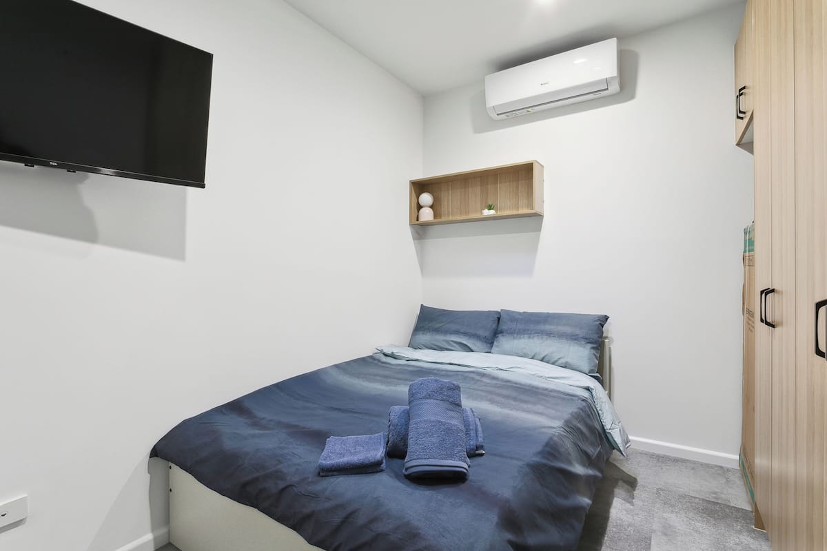 A bedroom with a bed, a flat-screen TV mounted on the wall, a wooden shelf with decorative items, and a wardrobe.