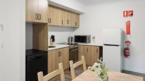 A compact kitchen with wooden cabinets, a microwave, a stove, and a refrigerator. There is a dining table with chairs and a vase of flowers. - Cozy Stays - Private Rooms (Adelaide)